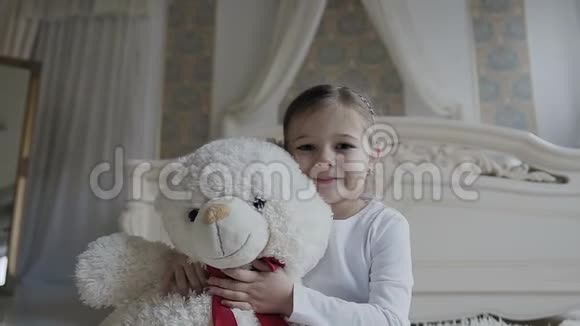 小女孩坐在床上的白色地毯上抱着一个柔软的熊玩具美丽的女孩和她的白色软玩具熊视频的预览图