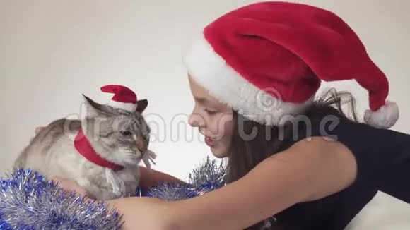 美丽的少女和她的猫戴着圣诞老人的帽子在白色背景的股票视频中快乐地接吻视频的预览图