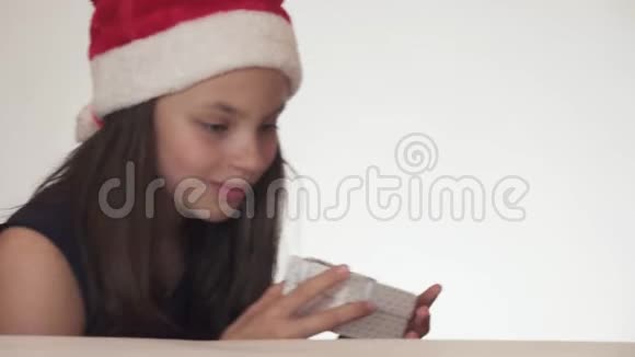 戴着圣诞老人帽子的漂亮淘气的少女偷偷地在桌子上放了一份白色背景的礼物视频的预览图