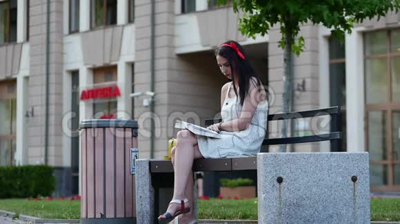一个头上有红色边缘的漂亮女孩坐在公园的长凳上看书在背景中你可以看到一个人是如何视频的预览图