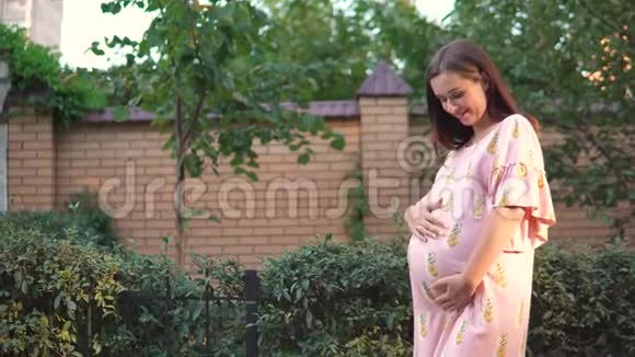 美丽的怀孕女孩穿着粉红色的裙子抚摸她的肚子在日落视频的预览图