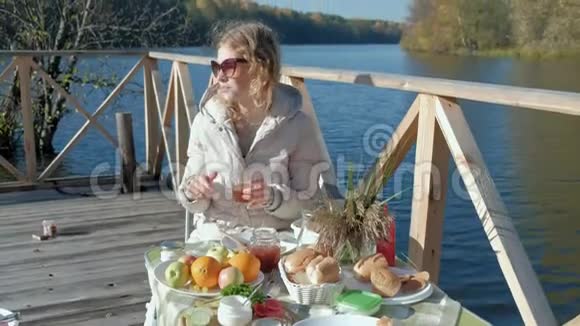 年轻女子戴着温暖的太阳镜坐在桌子旁喝着茶吃着带果酱的煎饼在河边野餐视频的预览图