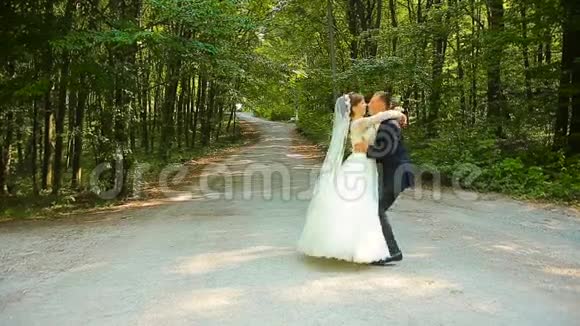 快乐的新郎和美丽的新娘穿着白色的裙子在绿色的森林里跳舞视频的预览图