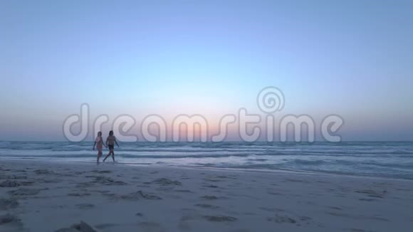这对年轻夫妇在黄昏时牵手在沙滩上散步穿比基尼的快乐女人和男朋友走在附近视频的预览图