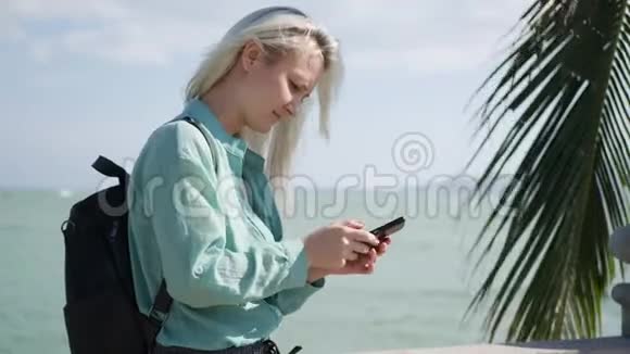 美丽的苗条女人留着长长的金发和绿色衬衫站在棕榈树附近在后台使用智能手机视频的预览图