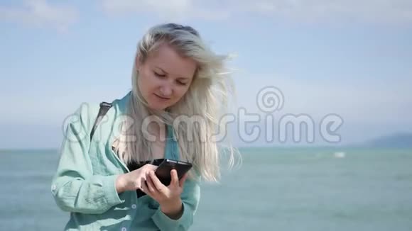 美丽的苗条女人长长的金发和绿色衬衫站立和使用智能手机在背景海女孩视频的预览图