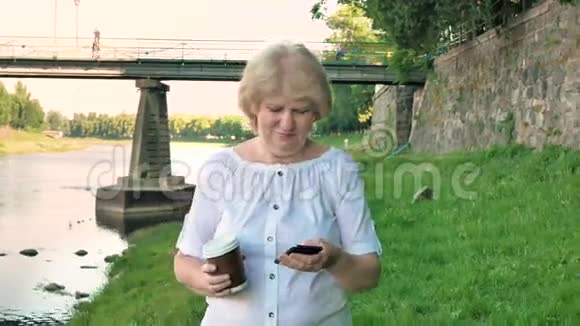 老年妇女使用智能手机和喝咖啡沿着海滨散步野石桥和老墙视频的预览图
