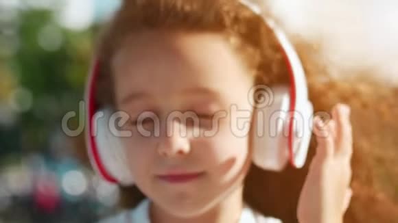 可爱的白种人卷发女孩穿着白色连衣裙坐在公园里戴着白色耳机听音乐看着里面的东西视频的预览图