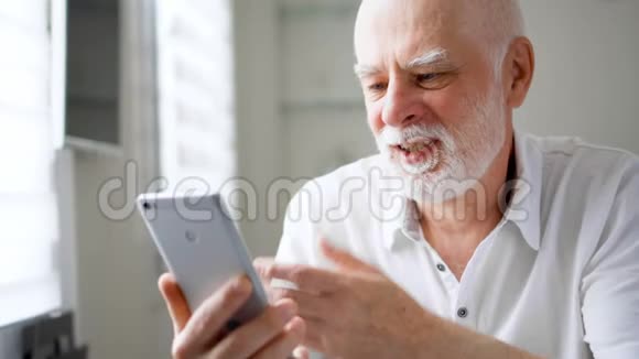 老人坐在家里拿着智能手机使用移动信使应用程序说话微笑着挥手视频的预览图