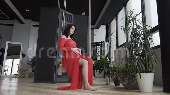 一个穿着红色长裙的怀孕女孩坐在秋千上轻轻地抚摸着她的腹部看着他概念视频的预览图