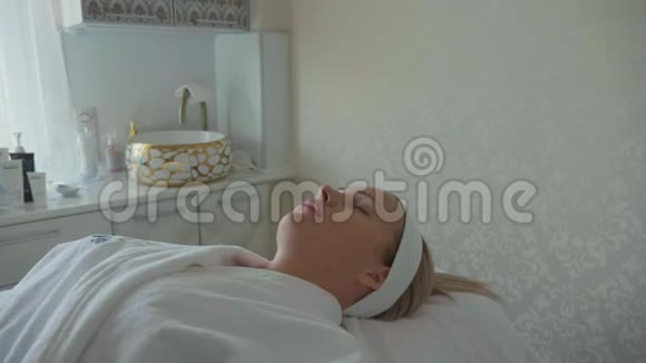美丽的女士穿着浴袍躺在水疗沙龙等待面部护理视频的预览图