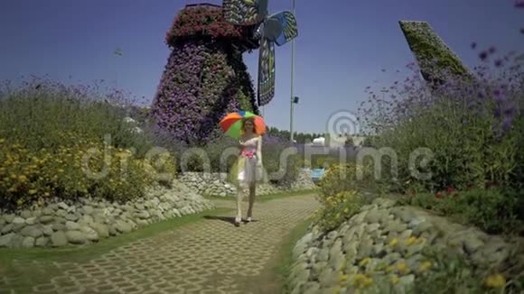 金发女郎带着彩色彩虹伞在花圃的小径上散步有趣的快乐梦可爱女孩视频的预览图