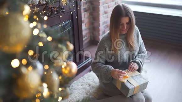 穿灰色毛衣的漂亮女人装饰礼物盒放在圣诞树下闪亮的圣诞树花环视频的预览图