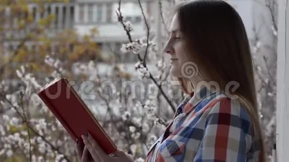 穿着格子衬衫的女孩站在窗边的阳台上读着一本关于盛开的杏树背景的书视频的预览图