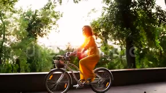 美丽的女人穿着黄色的长裙在黎明时骑着一辆城市自行车里面有一个篮子和花镜头耀斑视频的预览图
