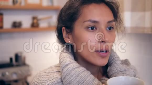 在寒冷的秋日年轻的白人女孩在温暖的毛衣里拿着杯子在厨房里喝热茶视频的预览图
