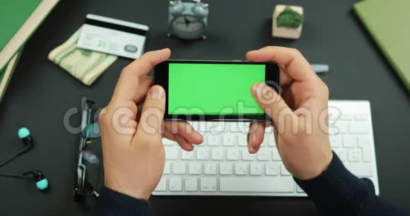 男人拿着一个绿色屏幕的黑色智能手机在工作桌上滚动一些东西视频的预览图
