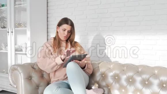 年轻女性坐在沙发上在平板电脑上浏览互联网在线支付在线帮助方便使用视频的预览图