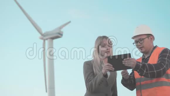 建筑业商务女士和年轻人戴着头盔和反光背心在风车背景使用数字视频的预览图