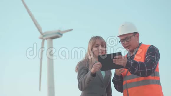 建筑业商务女士和年轻人戴着头盔和反光背心在风车背景使用数字视频的预览图