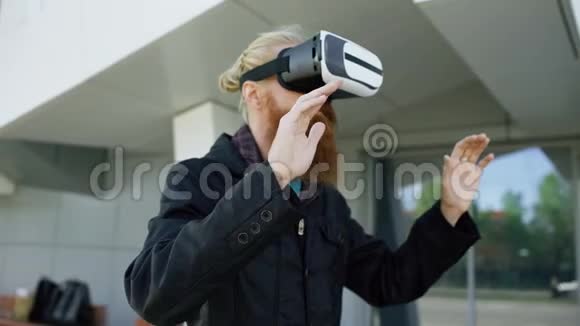 使用虚拟现实耳机进行360VR体验并带着微笑在户外的眼镜为年轻大胡子男子提供多莉镜头视频的预览图