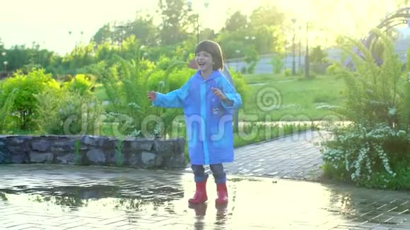 快乐可爱的小男孩喜欢跑步玩得开心穿着雨衣和橡胶靴在水坑里跳笑笑视频的预览图