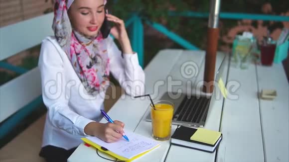 一个穆斯林妇女在手机上打电话在笔记本上做笔记视频的预览图