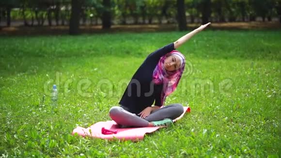 一个夏天戴着围巾的年轻穆斯林妇女在公园里伸展脊柱视频的预览图