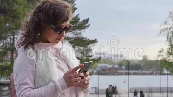 阳光明媚的一天戴着太阳镜的迷人年轻女子在公园散步时读写信息慢动作视频的预览图