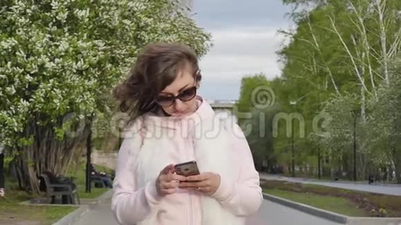 阳光明媚的一天戴着太阳镜的迷人年轻女子在公园散步时读写信息慢动作视频的预览图