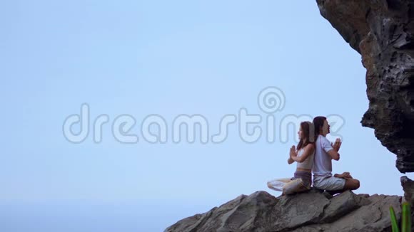男人和女人坐在山顶的岩石上背靠背冥想在大海的背景下做瑜伽视频的预览图