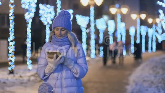 穿着冬装的金发美女在夜间灯光背景下使用平板电脑视频的预览图