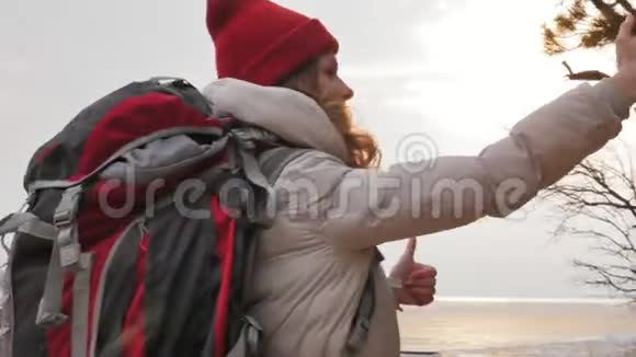 早春一位戴着红帽子的漂亮年轻女子背着一个大旅游背包走过森林带着一个小女孩视频的预览图