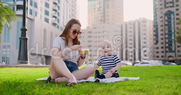 年轻的妈妈带着孩子坐在公园的草地上吃午饭视频的预览图