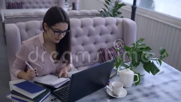 在网上学习漂亮的女孩用钢笔在笔记本上写笔记坐在桌子上拿着笔记本在咖啡馆里视频的预览图