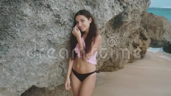 慢动作肖像美丽的快乐女孩穿着泳装在海滩上的岩石附近的波浪稳定射击视频的预览图