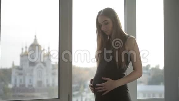 年轻的孕妇站在窗边抱着肚子视频的预览图
