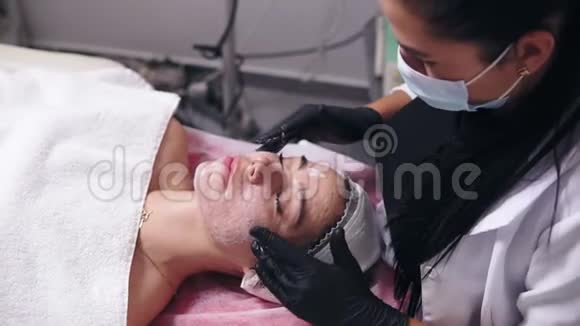 年轻女性皮肤有瑕疵躺在沙发上而专业的美容师正在涂面霜和做脸视频的预览图