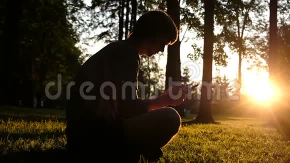 日落时分一个人手拿智能手机坐在草地上的剪影视频的预览图