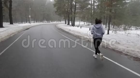 年轻迷人的白种人女孩在冬天带着耳机在雪地公园跑步后面跟着射击慢动作视频的预览图