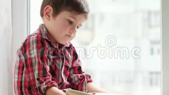 男孩翻着书页穿着衬衫的小孩坐在窗前拿着橘色封面的旧书视频的预览图