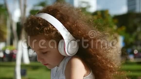 可爱的白种人卷发女孩穿着白色连衣裙坐在公园里戴着白色耳机听音乐看着里面的东西视频的预览图
