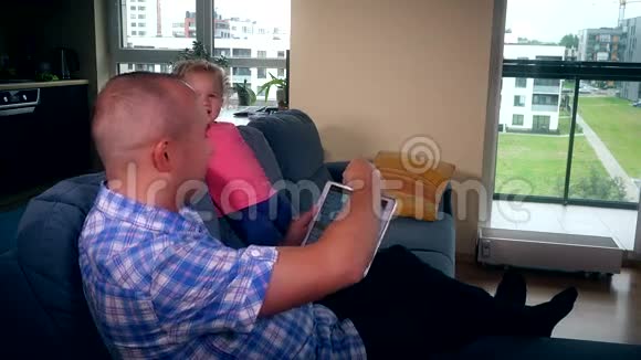 父亲在平板电脑上打开音乐快乐的活跃女婴在沙发上跳舞视频的预览图