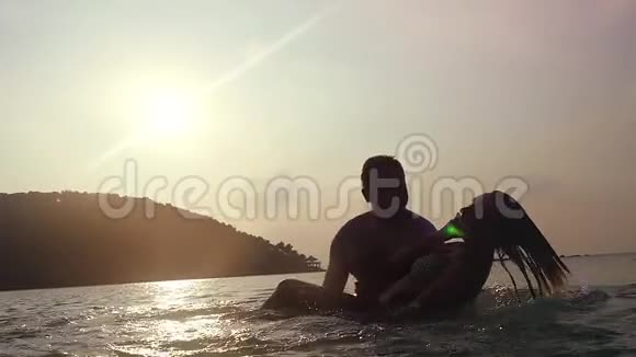 快乐的一对在海里度假男人抱着她的女孩在水里转悠大海在夕阳下飞溅视频的预览图