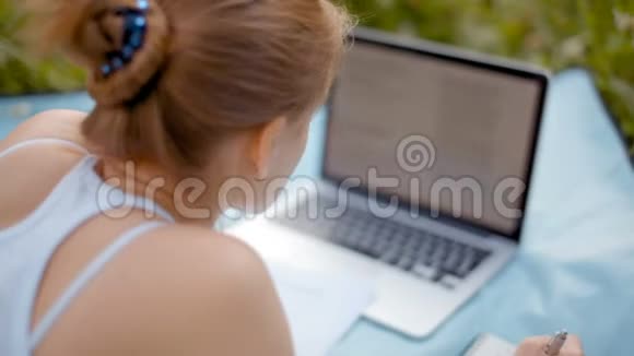 年轻的女学生夏天躺在公园里拿着笔记本和笔记本视频的预览图