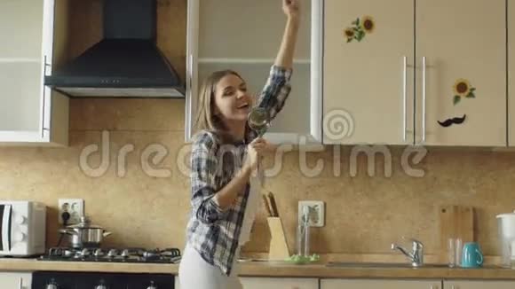 在厨房做饭的时候欢快的年轻风趣的女人一边用勺子跳舞一边唱歌视频的预览图