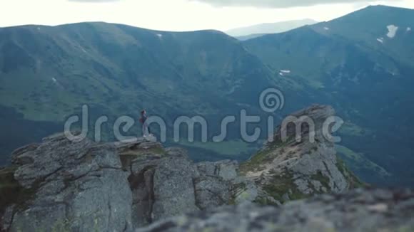 一位带背包的冒险女摄影师站在高高的岩石悬崖上俯瞰绿色的远处景色视频的预览图