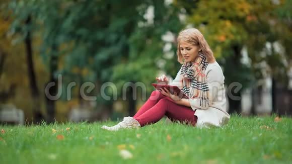 年轻女子在户外使用平板电脑坐在草地上微笑视频的预览图