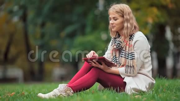 年轻女子在户外使用平板电脑坐在草地上微笑视频的预览图