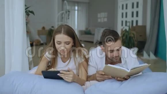 年轻夫妇有闲暇时间躺在床上在家里看书和平板电脑视频的预览图